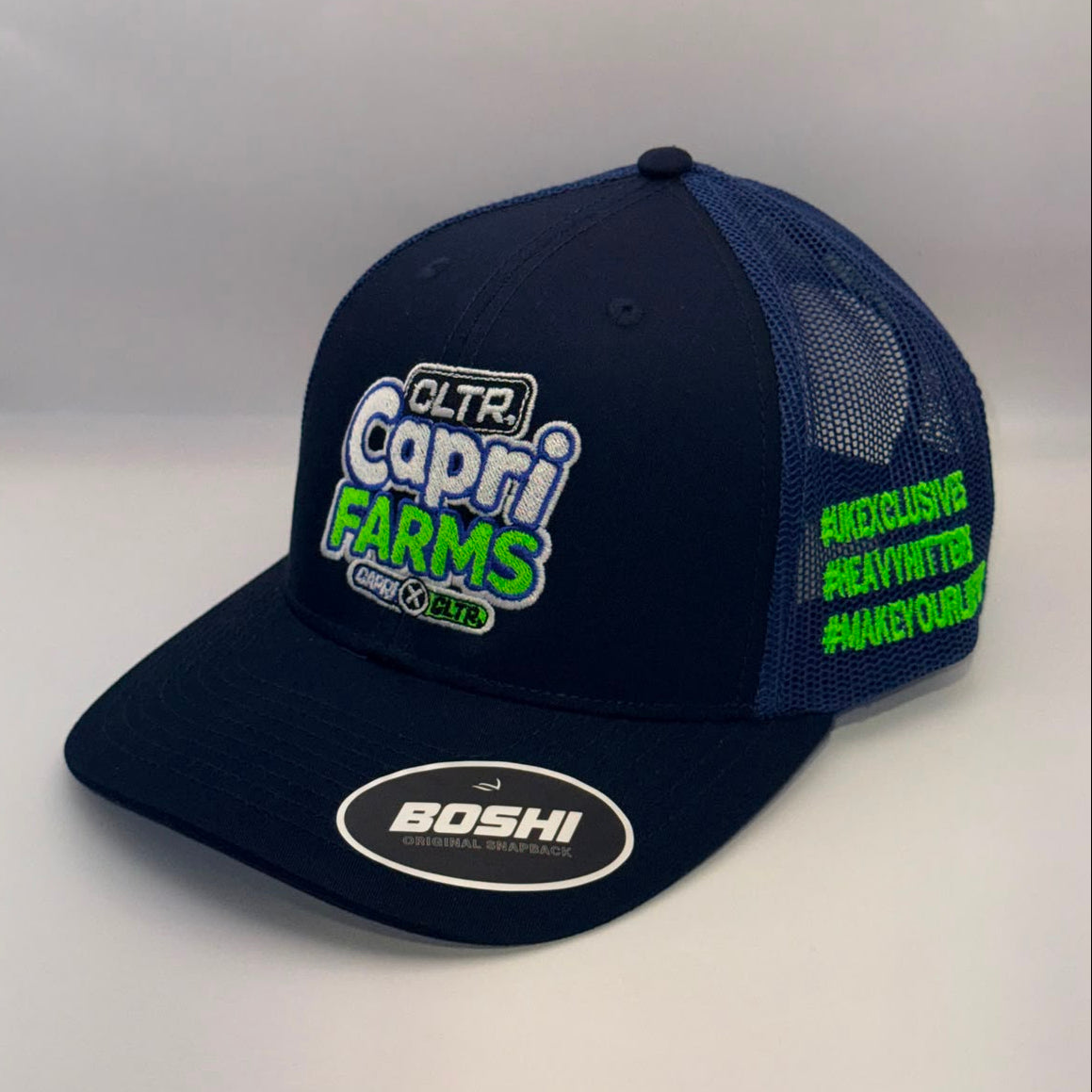 A navy blue trucker hat with a flat brim and mesh back, featuring a 'Capri Farms' logo on the front and neon green text on the side, with a 'BOSHI' sticker on the brim.